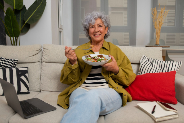 Mulher sentada em um sofá sorrindo e segurando um prato branco com comida.