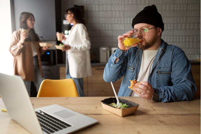 Homem sentado à mesa bebendo suco com uma marmita e laptop à sua frente. Atrás, duas mulheres conversam em pé segurando marmitas e copo.