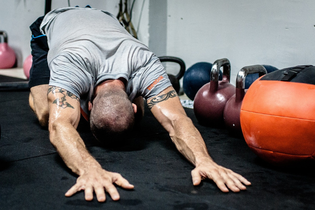 Um homem com camiseta cinza e tatuagens nos braços realiza um alongamento no chão de uma sala de ginástica. Ele está com a cabeça abaixada, tocando o chão com a testa e estendendo os braços para frente, numa postura que indica o término de um exercício ou uma pausa para recuperação. Ao fundo, podem-se ver diversos kettlebells coloridos e um grande medicine ball laranja, equipamentos típicos de treinamento funcional e CrossFit.