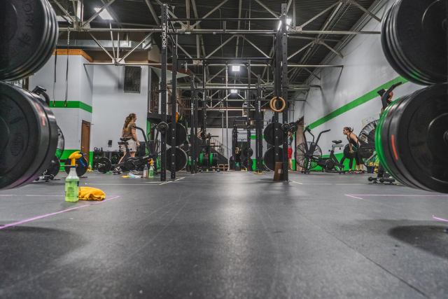 Uma imagem de uma academia de CrossFit capturada do nível do chão. Vemos barras olímpicas e discos de peso em primeiro plano, com espaço para treinamento funcional ao fundo. A academia apresenta um design industrial, com estruturas de aço, uma faixa verde nas paredes e iluminação suspensa. Pessoas estão espalhadas pelo espaço, algumas preparando-se para o treino e outras já engajadas em suas atividades físicas. Garrafas de água e toalhas estão visíveis no chão, sugerindo um ambiente de treino intenso.