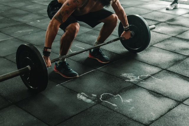 Um homem com tatuagens nos braços está agachado e prestes a levantar uma barra de peso em um piso emborrachado de academia, onde há marcas de magnésio deixadas por outras pessoas que treinaram antes dele. Ele usa tênis esportivos, meias pretas de cano alto, bermuda escura e uma pulseira esportiva. O ambiente sugere um treino intenso, provavelmente em uma sessão de CrossFit ou musculação.