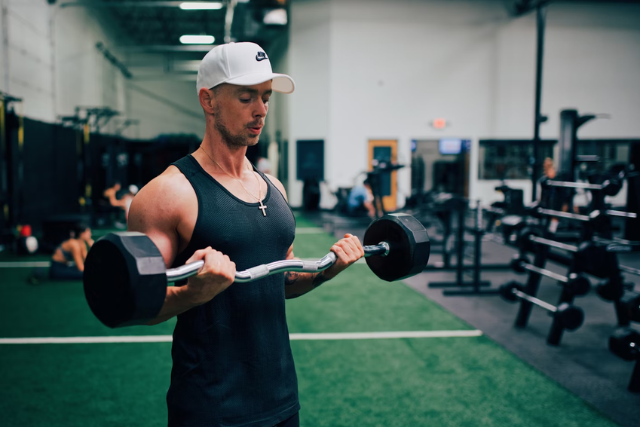 Um homem com uma regata preta e um boné branco está fazendo exercício com uma barra de pesos em uma academia. Ele está concentrado na execução do exercício de bíceps, conhecido como rosca direta. Ao fundo, pode-se observar um espaço amplo com equipamentos de ginástica, pesos livres e uma área de grama sintética, sugerindo um ambiente dedicado à prática de atividades físicas e musculação.